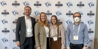 ICIS 2025 Gruppenfoto Das Bild zeigt eine Ganzkörperaufnahme von vier Personen, die nebeneinander vor einer Konferenz-Fotowand stehen und in die Kamera blicken. Der Hintergrund ist weiß und zeigt am oberen Rand in großer, dunkelblauer Schrift den Text „ICIS 2025“ sowie darunter „Nashville Tennessee December 14 – 17“. Flankiert wird der Text von grafischen Darstellungen einer Gitarre (links der Kopf, rechts der Hals in Orange mit dem Hashtag #ICIS2025). Der Rest der Wand ist mit einem wiederkehrenden Muster aus den Logos der „AIS“ (Association for Information Systems) und „ICIS 2025“ bedruckt. Die Personen werden von links nach rechts beschrieben: Ganz links steht ein Mann mit kurzen blonden Haaren und Brille. Er trägt ein dunkelgraues Sakko über einem weißen Hemd, dazu eine dunkelgraue Anzughose und braune Lederschuhe. Neben ihm steht eine Frau mit langen blonden Haaren, gekleidet in einen beigen, fein karierten Blazer, ein weißes Oberteil, eine weite schwarze Hose und helle Sneaker mit dunkler Sohle. Rechts daneben steht eine weitere Frau mit langen braunen Haaren und einer Brille. Sie trägt eine vertikal gestreifte Bluse in Weiß und Rotbraun, einen braunen, wadenlangen Rock, dunkle Strumpfhosen und hellbraune Stiefel mit auffälligem Fellbesatz. Ganz rechts steht ein Mann mit kurzen Haaren und Brille, der eine weiße FFP2-Maske trägt. Er ist mit einem blau-weiß karierten Hemd, einer dunklen Jeans und braunen Stiefeln bekleidet. Alle vier Personen tragen Konferenzausweise an weißen Bändern um den Hals. Sie stehen auf einem Teppichboden, der mit großen, abstrakten Blattmustern in Blau- und Grüntönen auf beigem Grund versehen ist.